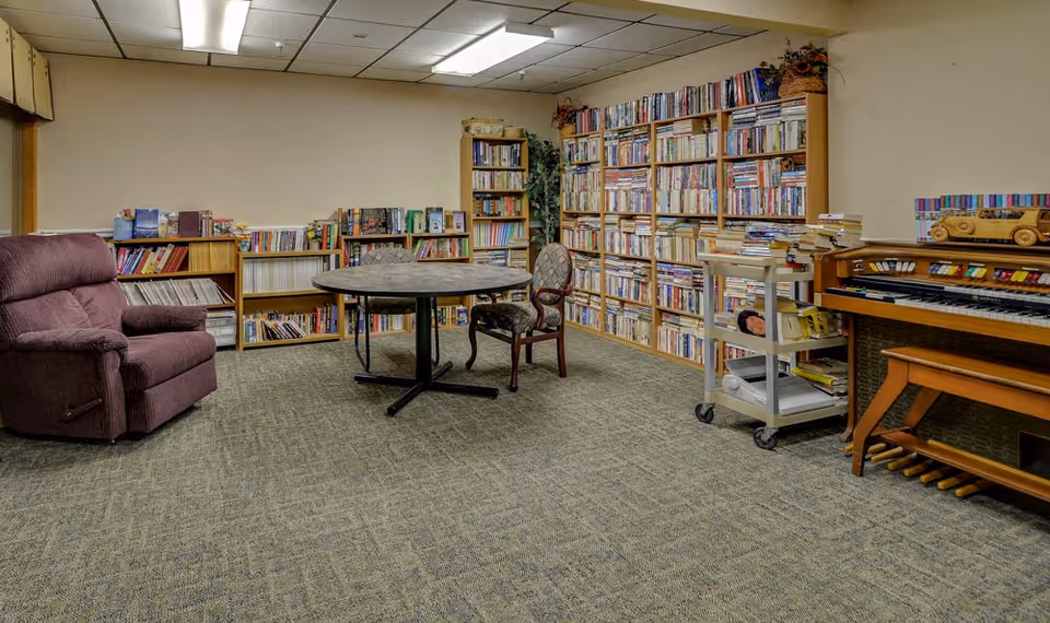 A cozy room with carpeted floor featuring a large bookshelf filled with books along the wall, a round table with two chairs, a purple recliner chair, a small rolling cart with books and items, and an organ with a wooden toy car on top.