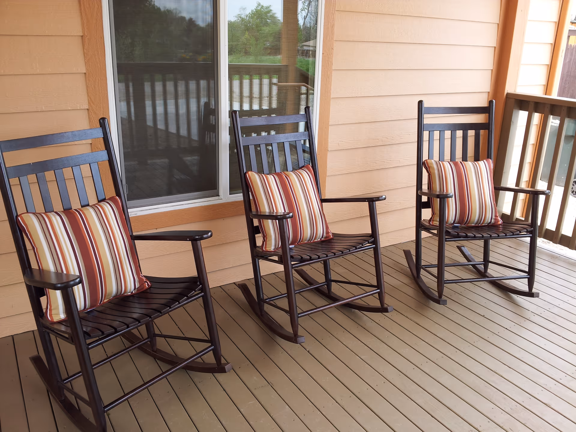Three dark wooden rocking chairs with striped cushions placed on a wooden porch with beige siding and a window in the background.