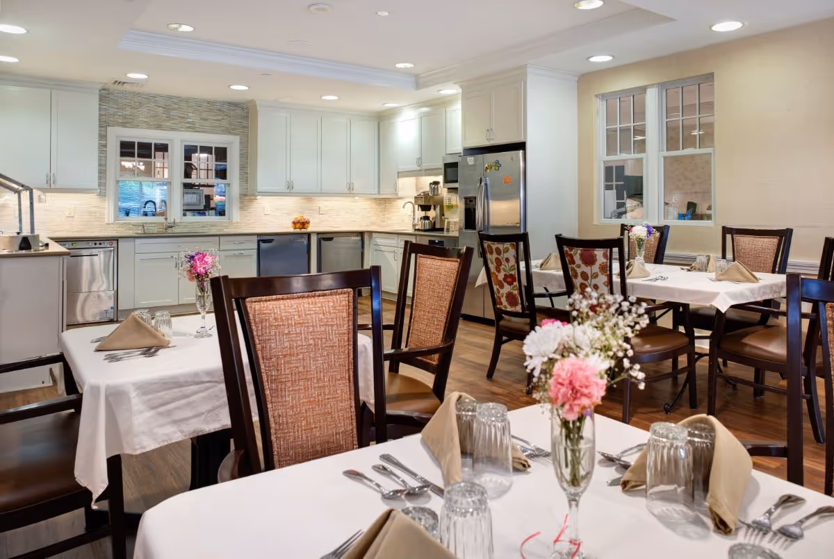 Bright dining room with neatly set tables and floral centerpieces in front of a white kitchen with stainless steel appliances.