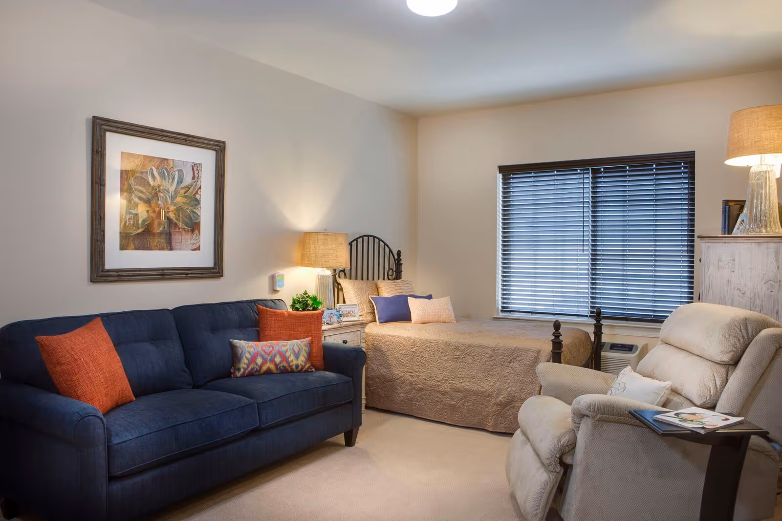 A cozy room with a beige single bed covered with a patterned quilt and several pillows, a dark blue sofa with colorful cushions, a beige recliner chair with a small side table holding magazines, two table lamps providing warm lighting, a framed floral artwork on the wall, and a window with closed blinds.