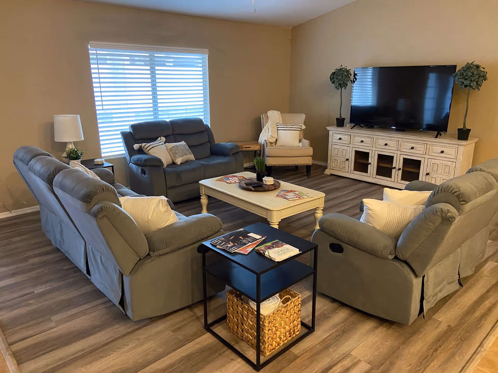 A cozy living room with beige walls and wood flooring, featuring two gray reclining sofas, a matching loveseat, a beige armchair with pillows, a white wooden coffee table with magazines and a small plant, a black side table with books and a woven basket underneath, a large flat-screen TV on a white wooden TV stand, and two potted plants on either side of the TV.