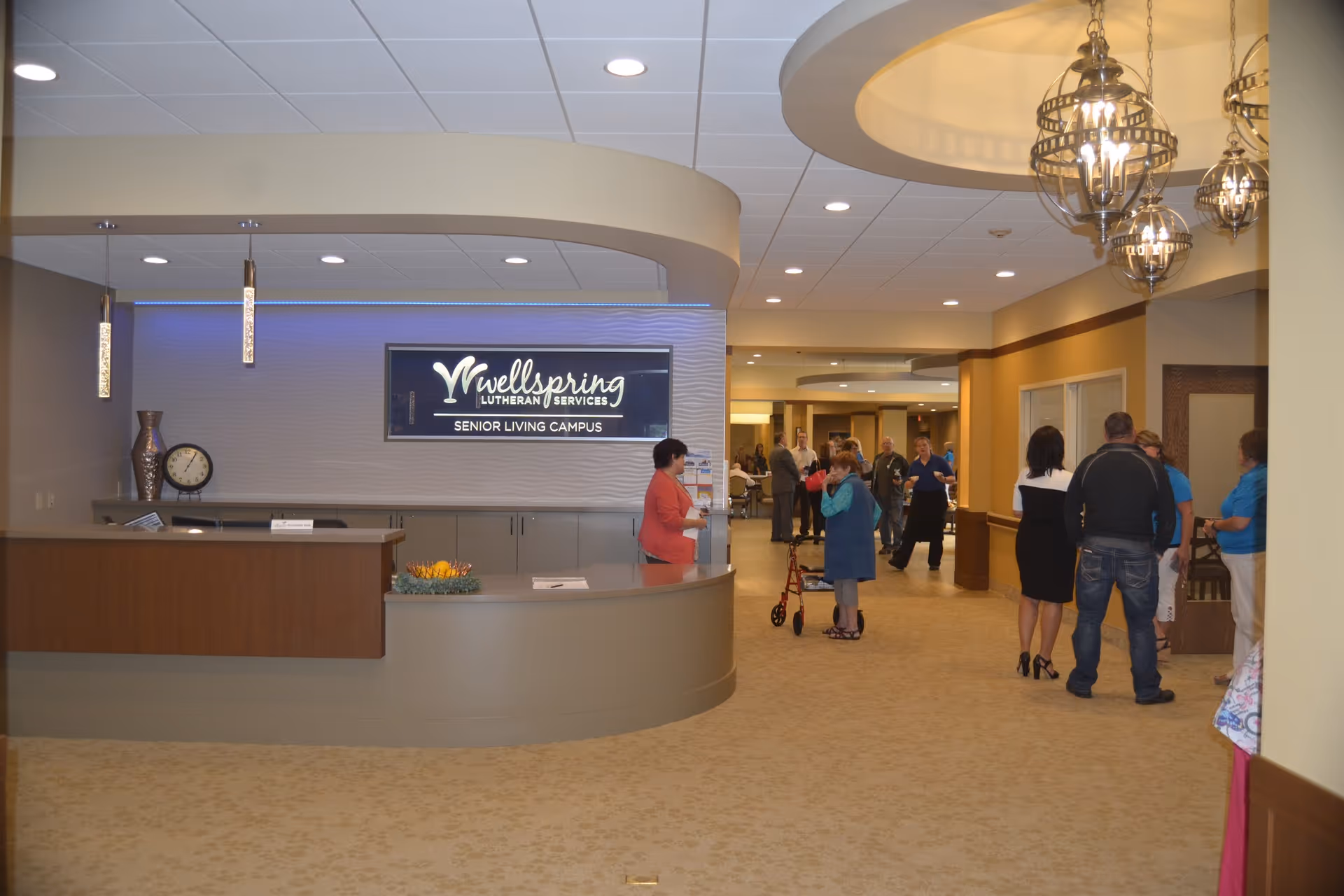 Reception area of Wellspring Lutheran Services Senior Living Campus with a curved front desk, decorative vases, a clock, and a sign on the wall. Several people are standing and conversing in the hallway beyond the reception desk.