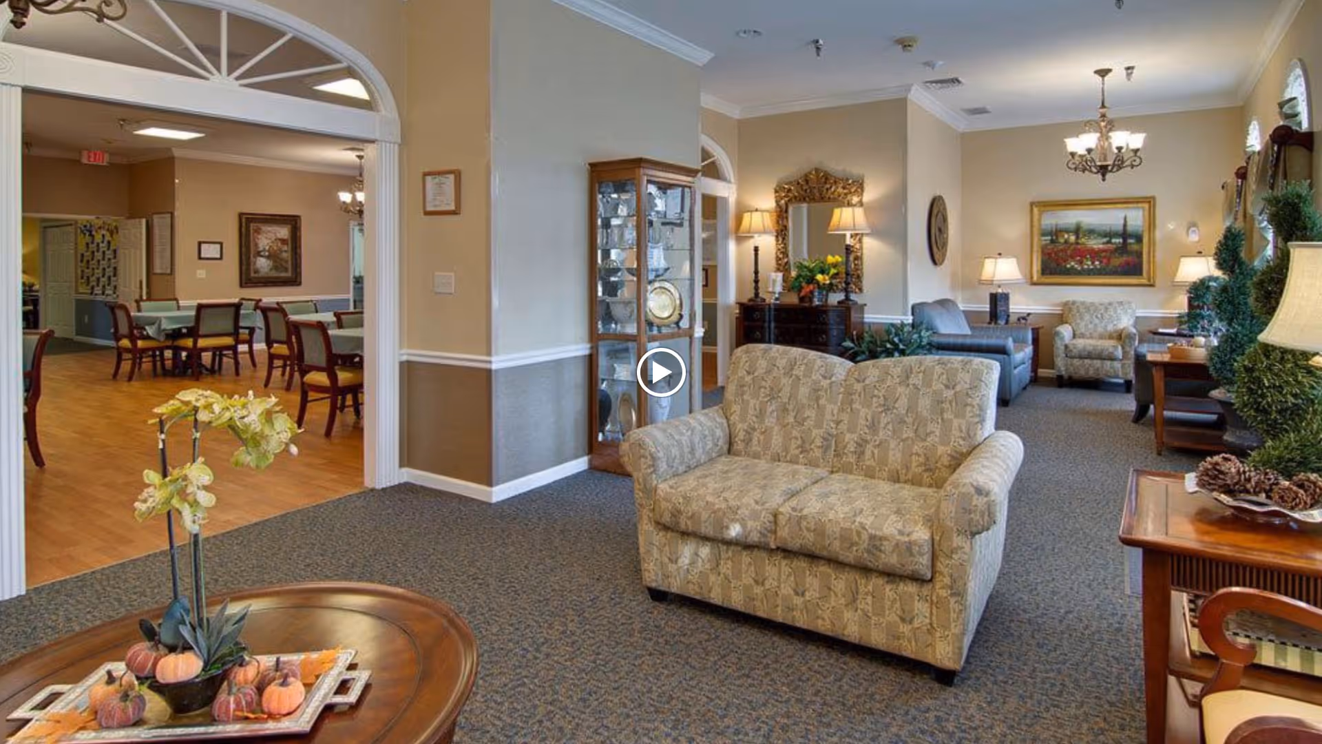 A cozy senior living facility common area with patterned loveseat, armchairs, wooden tables, lamps, and decorative plants. In the background, a dining area with multiple tables and chairs is visible through an arched doorway. The walls are painted beige with white trim, and the room is warmly lit with ceiling lights and table lamps.