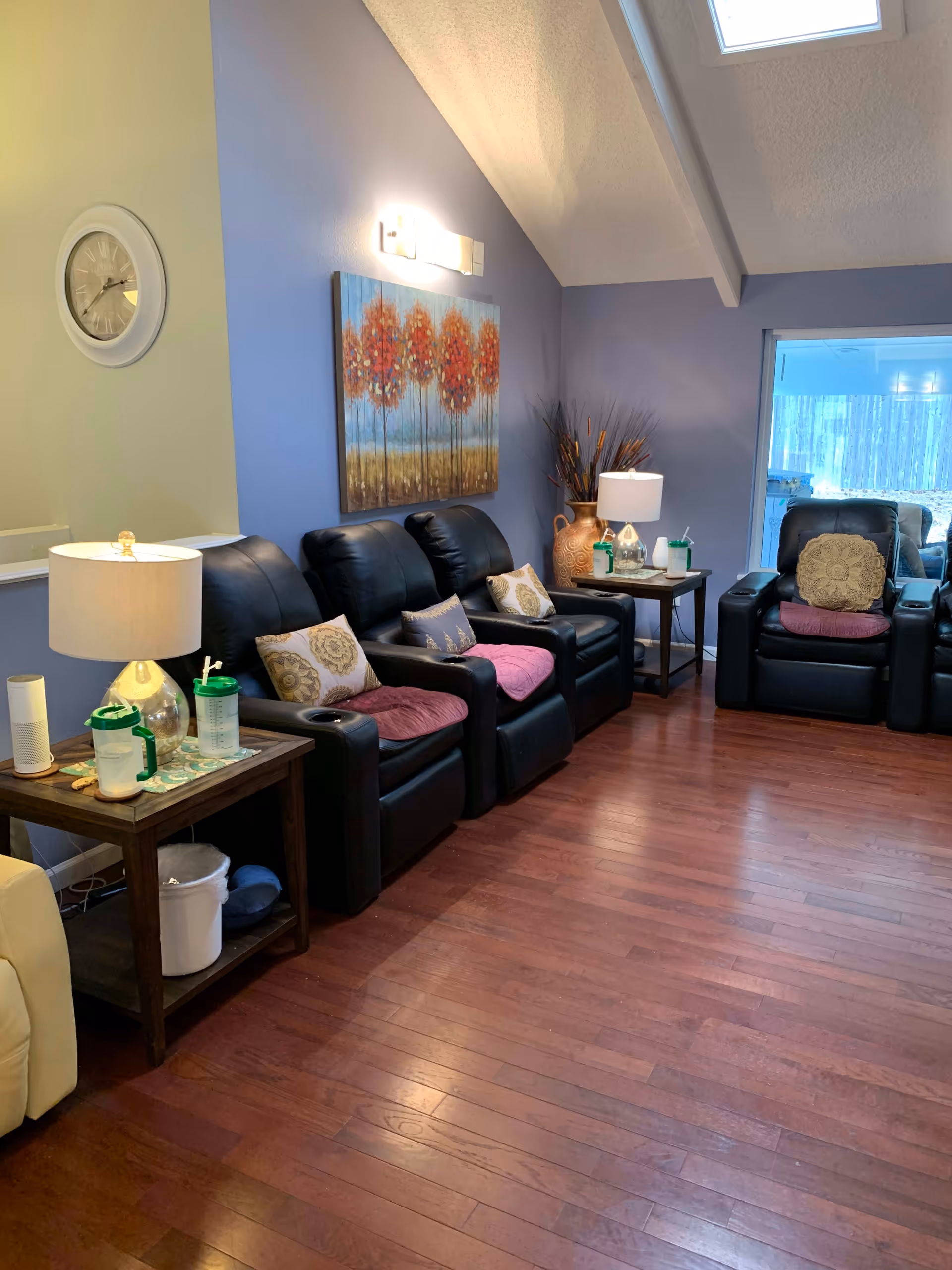 A cozy living room with three black leather recliners arranged side by side, each with decorative pillows and pink seat cushions. There are two wooden side tables with lamps, water bottles, and other small items. The walls are painted in light blue and beige, with a large painting of orange trees hanging above the recliners. The room has wooden flooring and a skylight on the ceiling, with a clock on the beige wall and a window showing an outdoor view.
