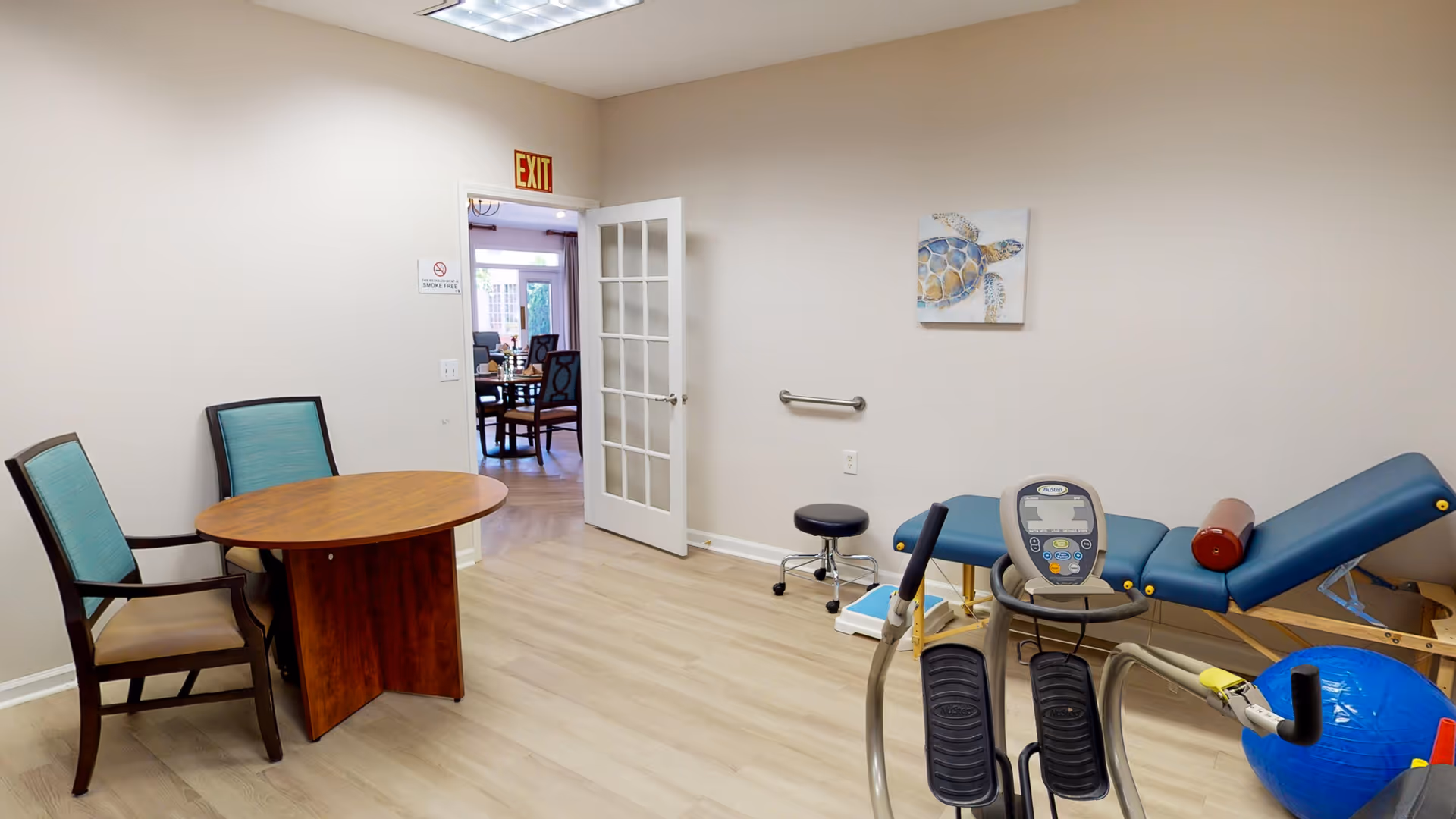 Interior therapy/exercise room with an elliptical machine, treatment table, round table and chairs, and an open door with an EXIT sign.