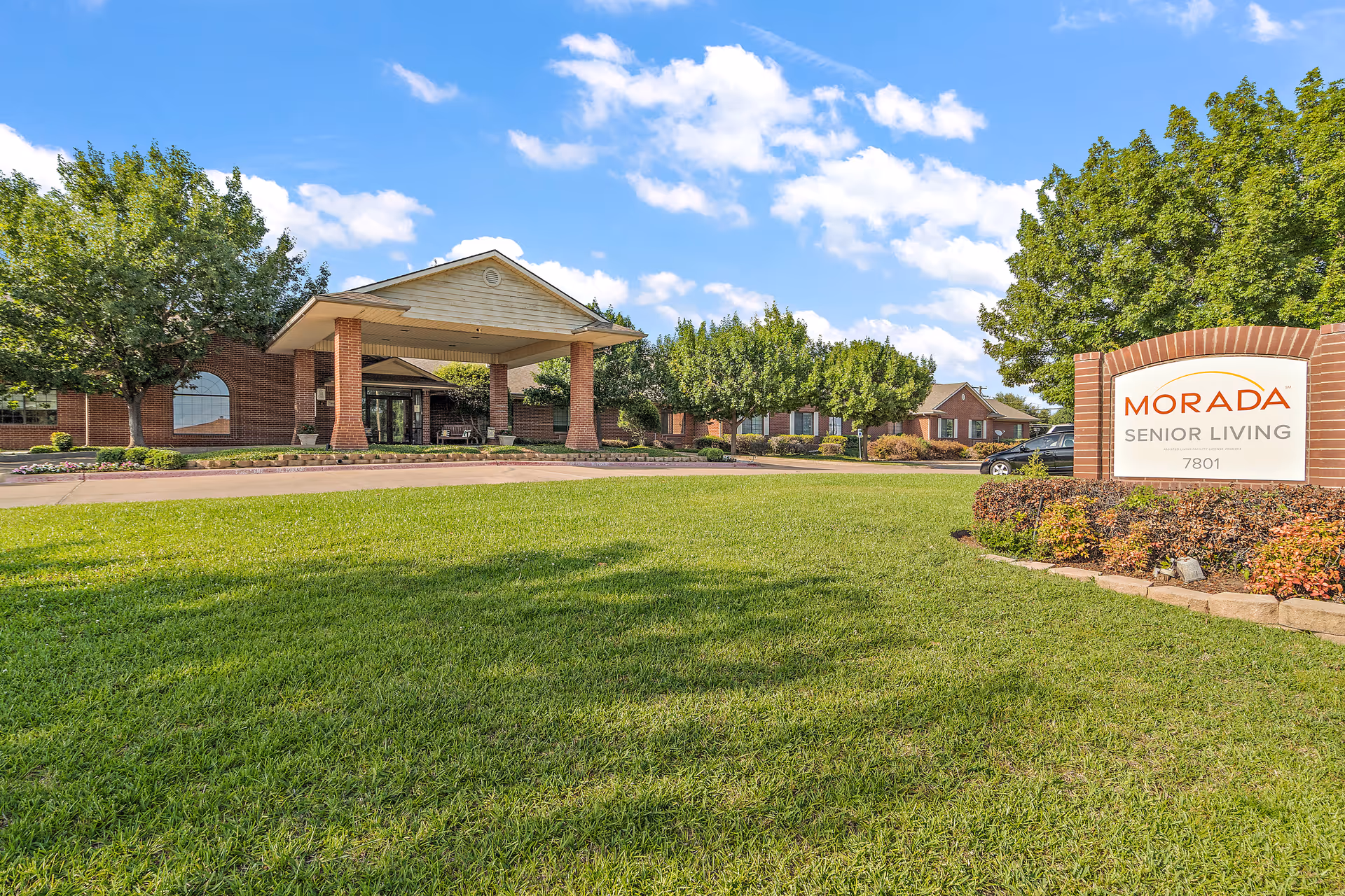 Exterior view of Morada Senior Living facility in North Richland Hills, showing a well-maintained lawn, trees, and a covered entrance with brick pillars under a blue sky with scattered clouds. A sign with the facility name and address is visible on the right side.