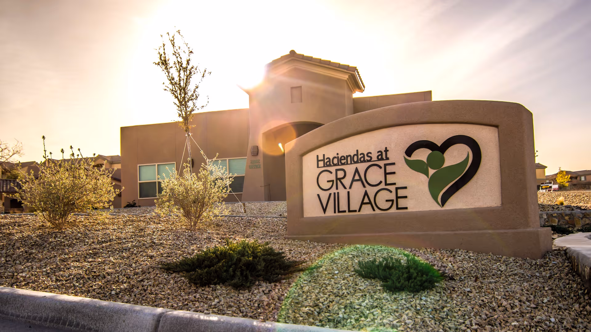 Exterior view of the Haciendas at Grace Village facility sign with a beige building and landscaping featuring small bushes and gravel under a bright sky with the sun shining.
