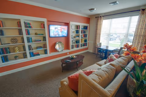 A cozy living room with a beige sofa adorned with red and patterned pillows, a wooden coffee table with decorative items, built-in white bookshelves filled with books and decor, a flat-screen TV mounted on a red accent wall, and a window with floral curtains letting in natural light. A small table and chairs are placed near the window.