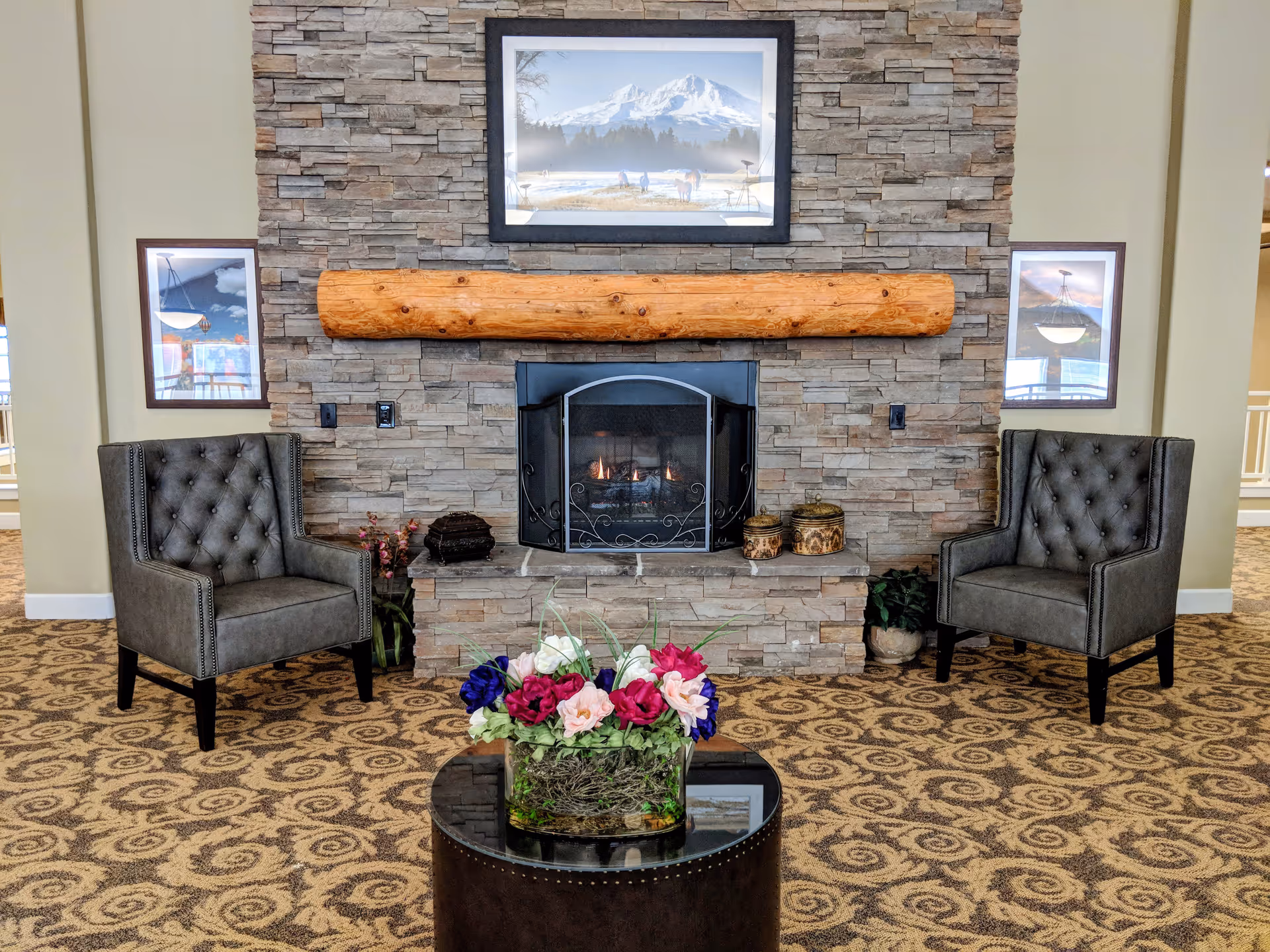 A cozy seating area in a senior living facility featuring a stone fireplace with a wooden mantel. Two gray tufted armchairs are placed on either side of the fireplace. A round black coffee table with a colorful floral arrangement sits in front of the fireplace. The walls have framed landscape pictures, and the carpet has a patterned design.
