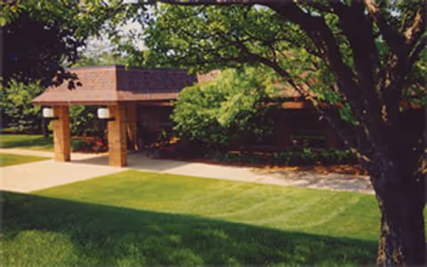 Exterior view of a single-story building with a covered entrance supported by brick columns, surrounded by green grass and trees.