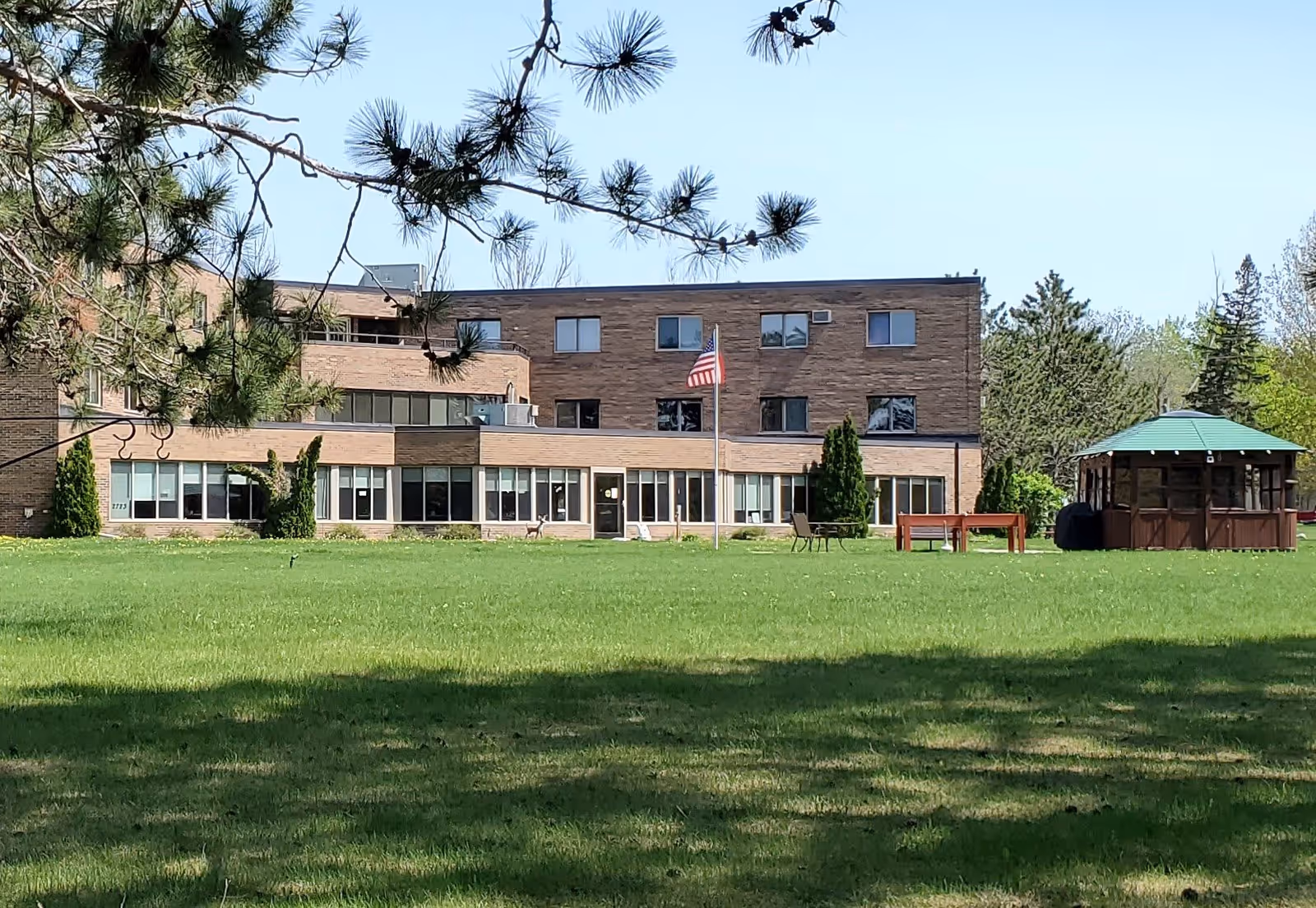 Exterior view of a multi-story brick building with large windows, an American flag on a flagpole in front, a gazebo with a green roof, outdoor chairs, and a table on a well-maintained grassy lawn with trees in the background.