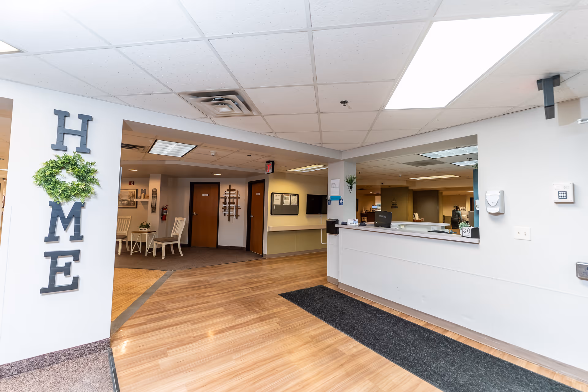 Reception area of a nursing and rehab facility with a front desk, waiting chairs, and a decorative 'HOME' sign on the wall.