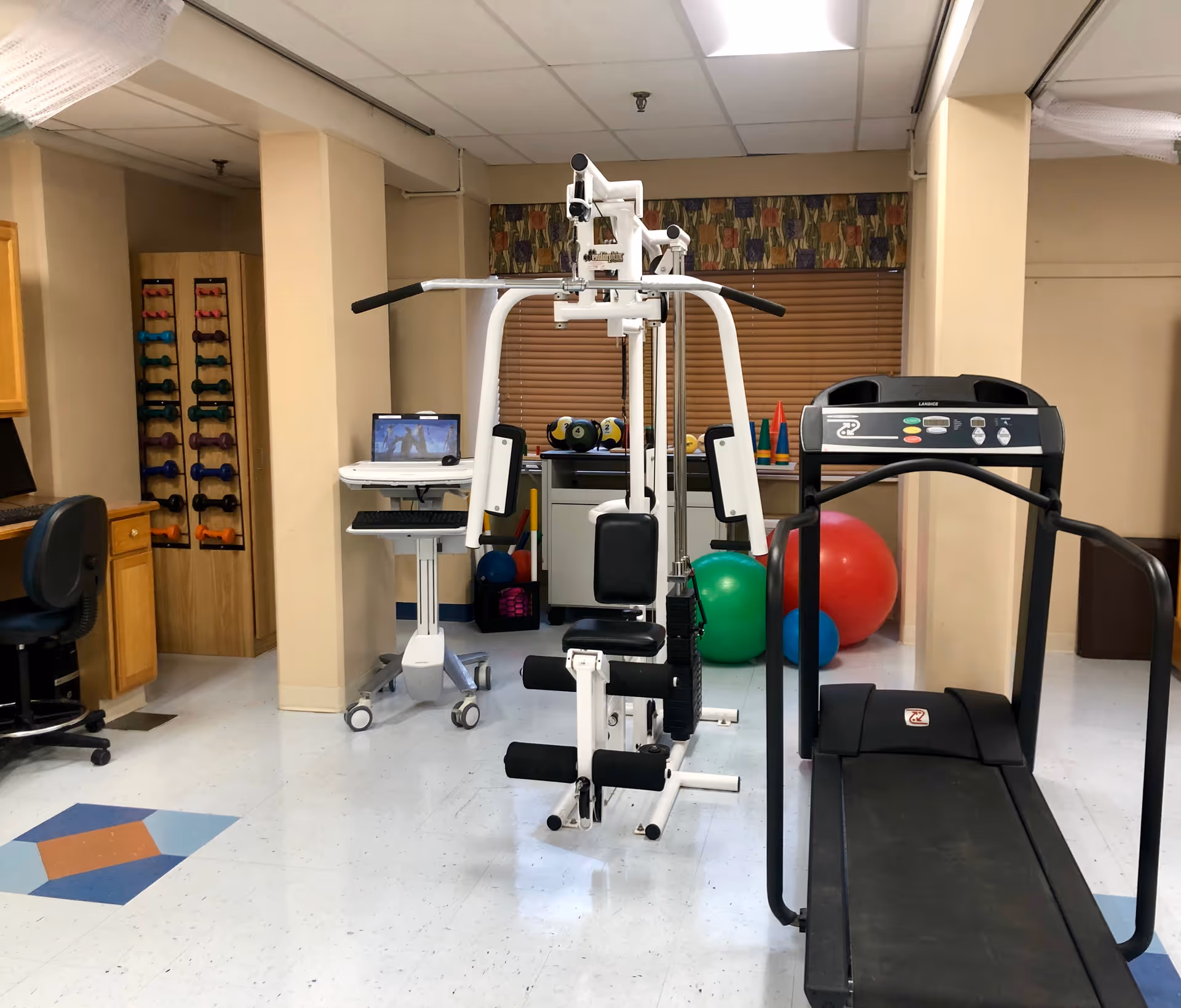 Interior view of a rehabilitation and health care center gym area featuring exercise equipment including a treadmill, a multi-function weight machine, colorful exercise balls, and a rack of dumbbells. There is also a computer workstation with a chair and a monitor on a mobile stand. The room has beige walls, a tiled floor with a blue and orange diamond pattern, and a window with a floral valance and closed blinds.