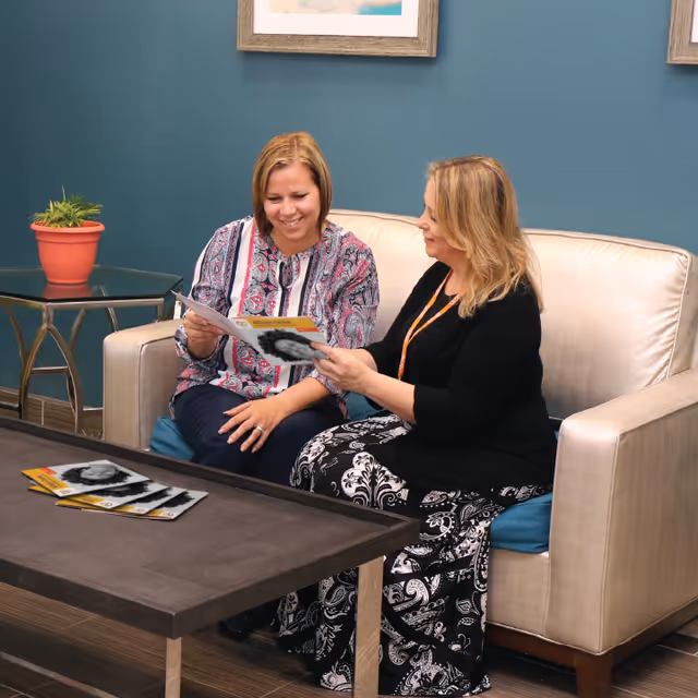 Two women sitting on a beige couch in a room with teal walls, looking at brochures and smiling. There is a dark wooden coffee table in front of them with more brochures on it, and a small side table with a potted plant next to the couch.