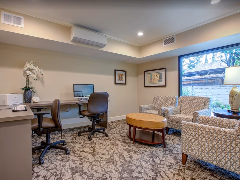 A cozy interior common room with desks and office chairs on the left and patterned armchairs around a coffee table by a large window.