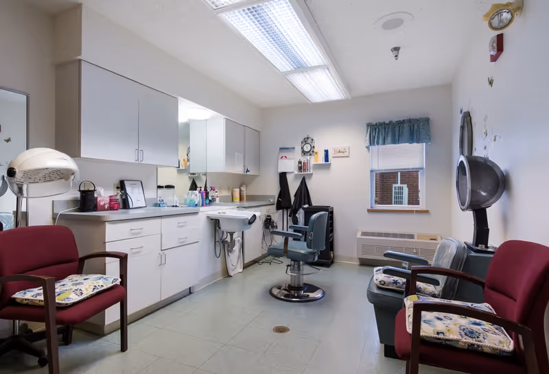 Interior of a hair salon room in a senior living facility with salon chairs, hair drying machines, a sink, cabinets, and a window with a blue valance.