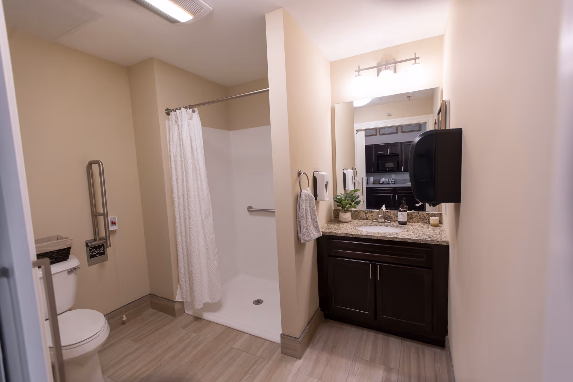 Accessible bathroom with a walk-in shower and curtain, toilet with grab bar, and a dark wood vanity with mirror and soap dispenser.