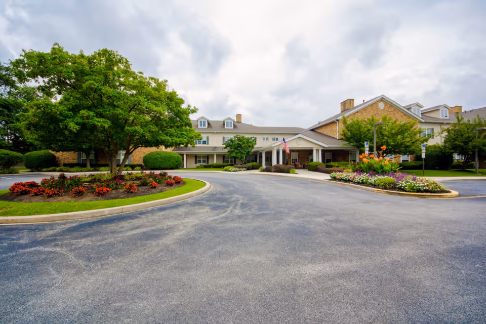 Front exterior view of Somerford House And Place Newark, showing a large building with beige brick and white siding, surrounded by landscaped flower beds, green trees, and a circular driveway under a cloudy sky.