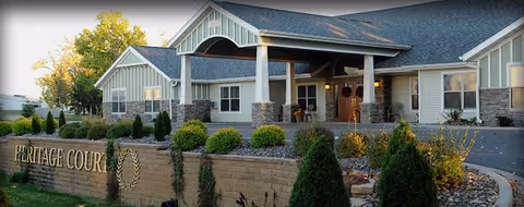 Exterior view of Heritage Court senior living facility showing a single-story building with a covered entrance, stone and siding facade, landscaped shrubs, and a brick sign with the facility name.