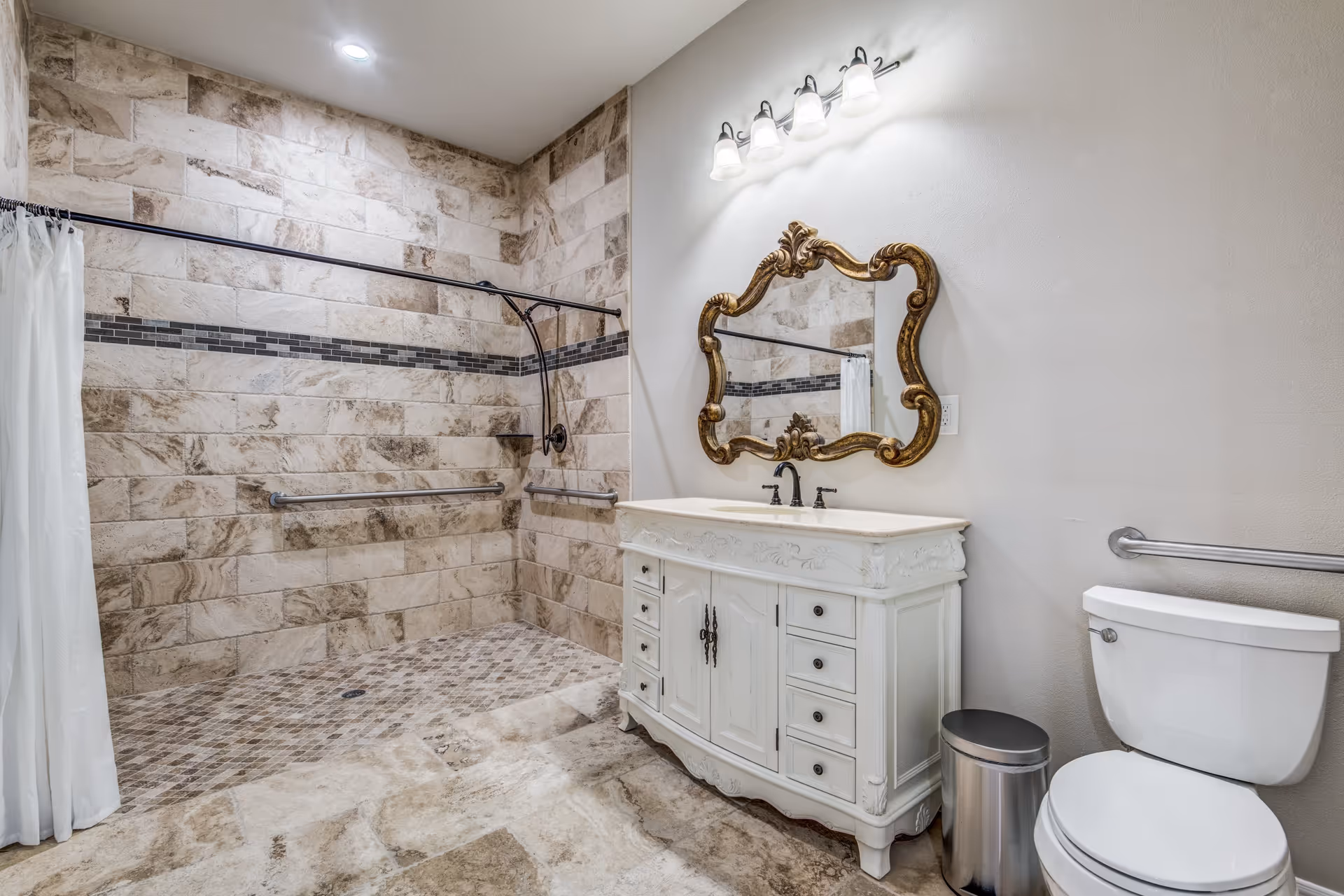 A spacious bathroom with beige and brown tiled walls and floor. The walk-in shower has a white curtain and two metal grab bars. There is a white vanity with ornate detailing and a matching decorative mirror above it. A toilet and a small metal trash can are positioned to the right of the vanity. The bathroom is well-lit with a row of five lights above the mirror.