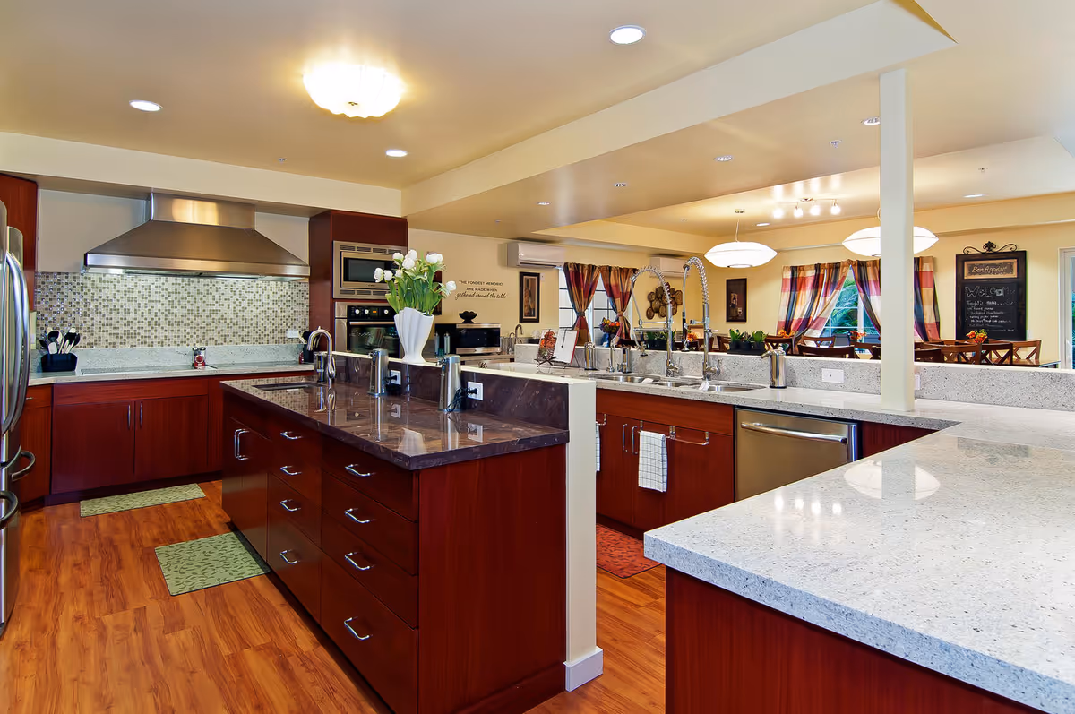 A spacious, modern kitchen with wooden cabinets, a large island with a dark countertop, stainless steel appliances including a refrigerator, oven, and range hood. The kitchen opens into a dining area with colorful curtains and wooden chairs around tables. The space is well-lit with ceiling lights and decorated with flowers and wall art.