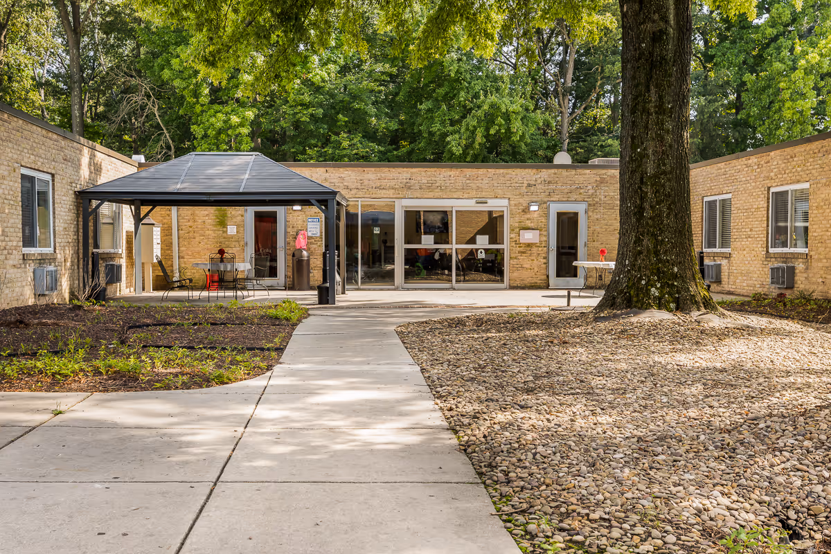 Outdoor courtyard area of Autumn Lake Healthcare at Baltimore Washington featuring a large tree surrounded by rocks, a paved walkway, a covered seating area with tables and chairs, and a brick building with multiple windows and doors in the background.