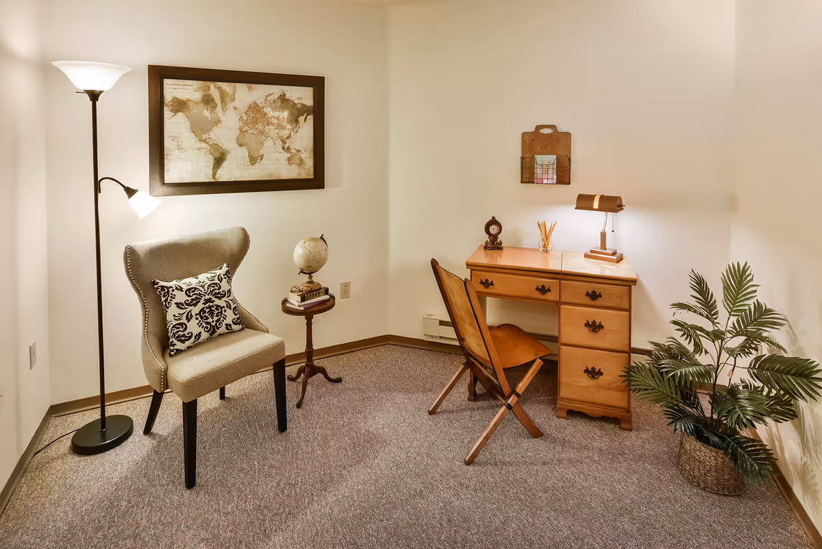 A cozy interior room with a beige upholstered chair featuring a decorative black and white pillow, a small round wooden side table with a globe and books, a wooden desk with drawers and a wooden chair, a desk lamp, a small clock, a reed diffuser, a wall-mounted wooden organizer, a tall floor lamp, a framed world map on the wall, and a potted plant in a woven basket on a carpeted floor.