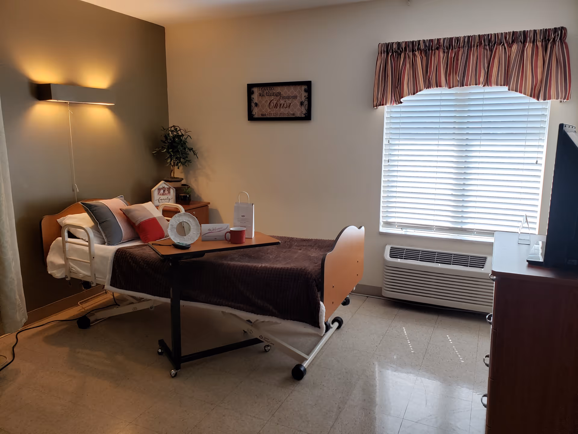 A senior living facility bedroom with a hospital-style bed covered with a brown blanket and several pillows. A small overbed table holds a red mug, a white decorative item, and a welcome card. There is a wooden nightstand with a small plant and decorative items next to the bed. A window with blinds and a striped valance lets in natural light. A wall-mounted light fixture illuminates the room softly. A dresser with a TV on top is visible on the right side of the room.