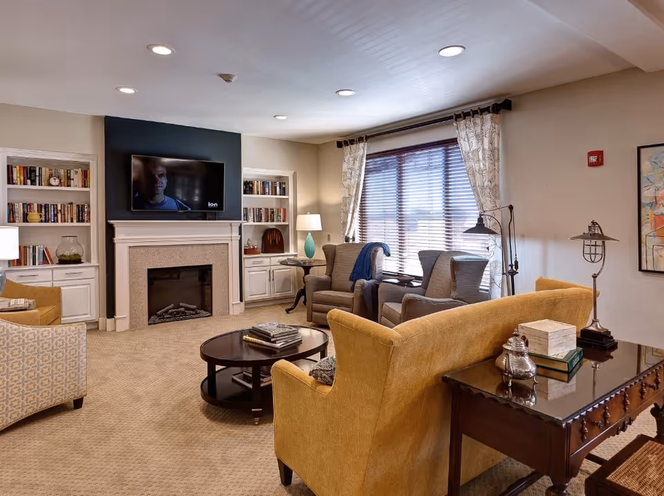 Well-appointed common living room with sofas and armchairs arranged around a coffee table, fireplace, wall-mounted TV and bookshelves.