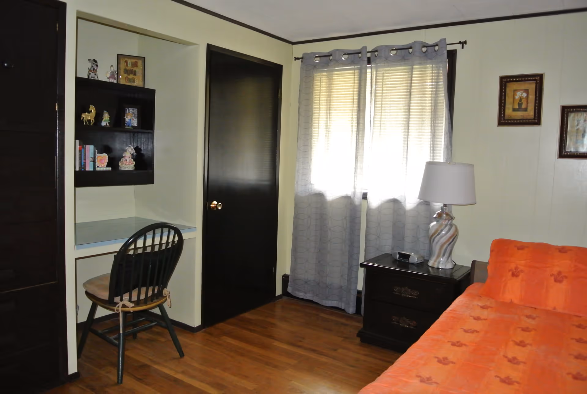 A bedroom with a wooden floor, an orange bedspread on the bed, a black nightstand with a decorative lamp and a clock, a window with sheer gray curtains, a black door, and a built-in desk area with shelves holding small decorative items and books. A wooden chair with a cushion is placed at the desk.