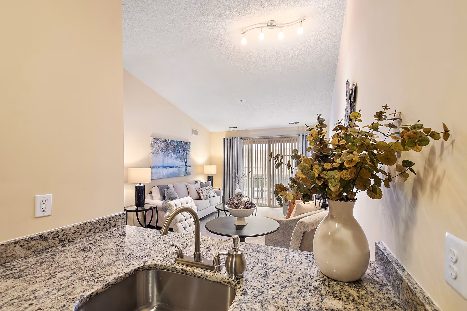View from a kitchen counter with a granite countertop and a stainless steel sink looking into a living room area. The living room has a beige sofa, a round coffee table with decorative items, two side tables with lamps, a large wall painting, and sliding glass doors with vertical blinds leading outside. A large white vase with green and brown foliage is placed on the kitchen counter.