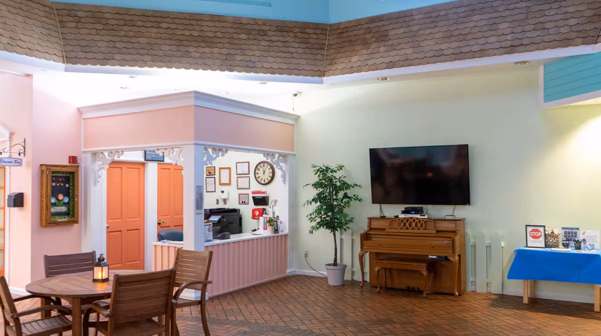 Interior common area of a senior living facility featuring a small reception desk with office equipment and framed certificates on the wall, a wooden piano with a flat-screen TV mounted above it, a potted plant, a round wooden table with four chairs, and a blue table with informational materials. The ceiling has a decorative shingle design and the floor is covered with brick-patterned tiles.