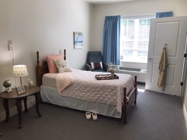 A neatly arranged bedroom with a single wooden bed covered in a light pink quilt and pillows. A small wooden side table with a lamp, a globe, and a framed photo is beside the bed. A pair of slippers is placed on the carpeted floor next to the bed. A blue armchair is positioned near a window with blue curtains, and the door to the room is open with a scarf hanging on the handle.