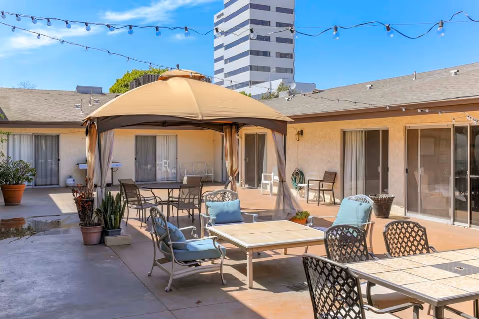 Outdoor courtyard area at Sea Cliff Assisted Living featuring a beige canopy with a table and chairs underneath, additional tables and chairs with blue cushions, potted plants, string lights overhead, and sliding glass doors leading to the interior rooms.