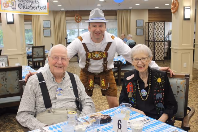 Two older adults seated at a decorated table with a man in lederhosen standing behind them during an Oktoberfest celebration in a dining room.