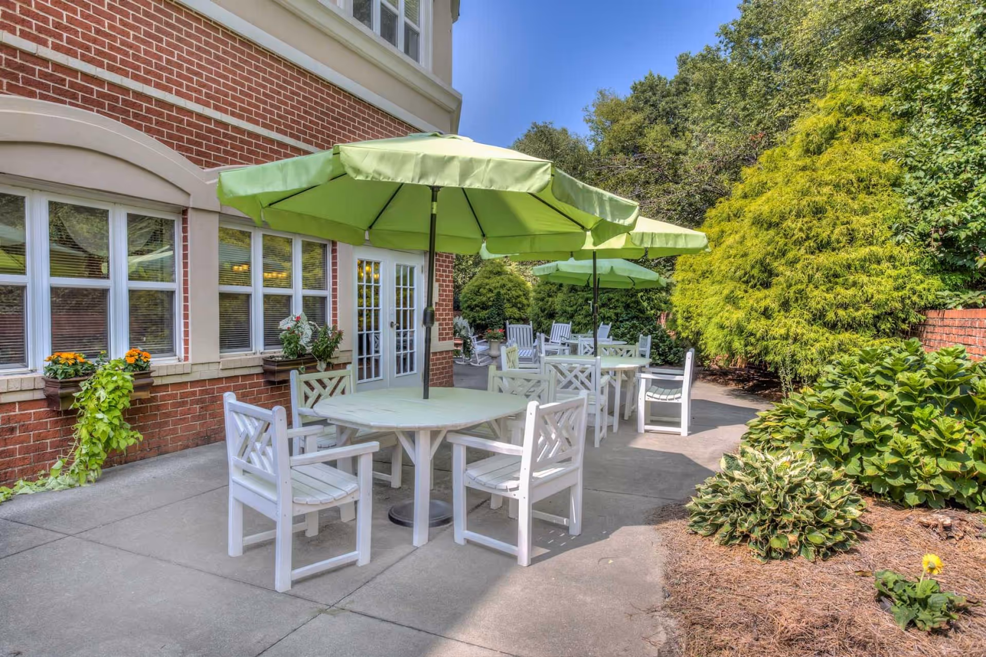Outdoor patio area with white wooden tables and chairs under green umbrellas next to a brick building with windows and French doors. The patio is surrounded by lush green bushes and trees under a clear blue sky.