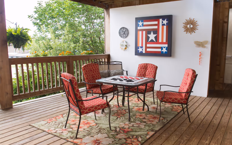 Covered wooden porch with red cushioned chairs around a table on a floral rug and patriotic wall art on the wall.