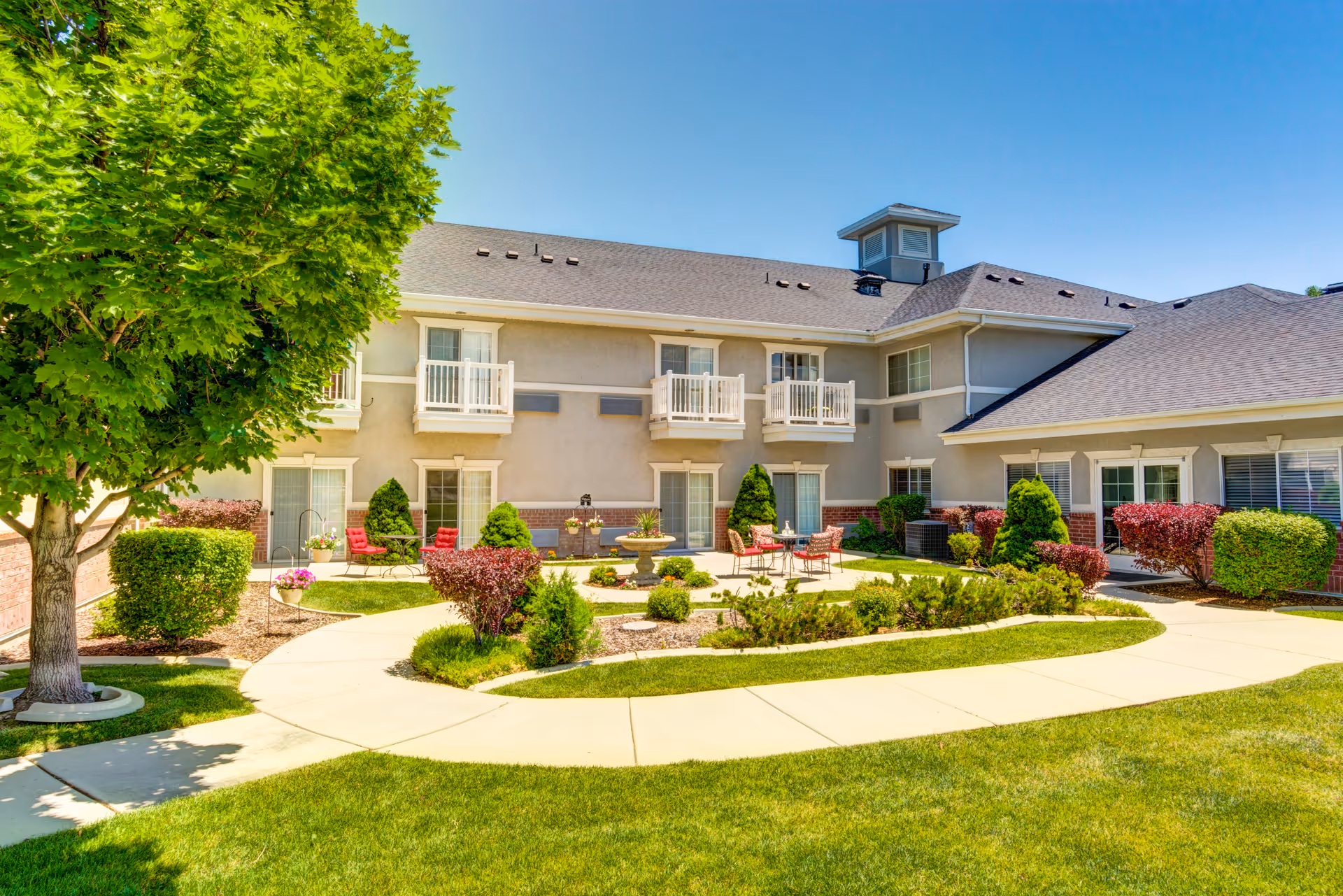 Outdoor courtyard area of a senior living facility with a well-maintained lawn, paved walkways, small trees, shrubs, and patio seating with tables and chairs. The building has two stories with balconies and large windows under a clear blue sky.