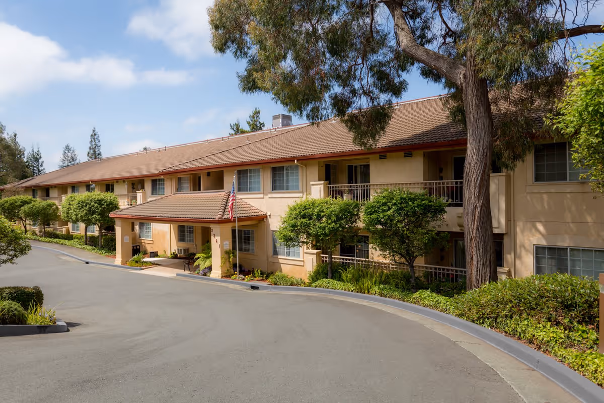Exterior view of a two-story senior living facility building with beige walls and a brown tiled roof. The entrance is covered with a small roof supported by columns, and an American flag is displayed near the entrance. The building is surrounded by well-maintained greenery including trees and bushes, with a paved driveway in front.