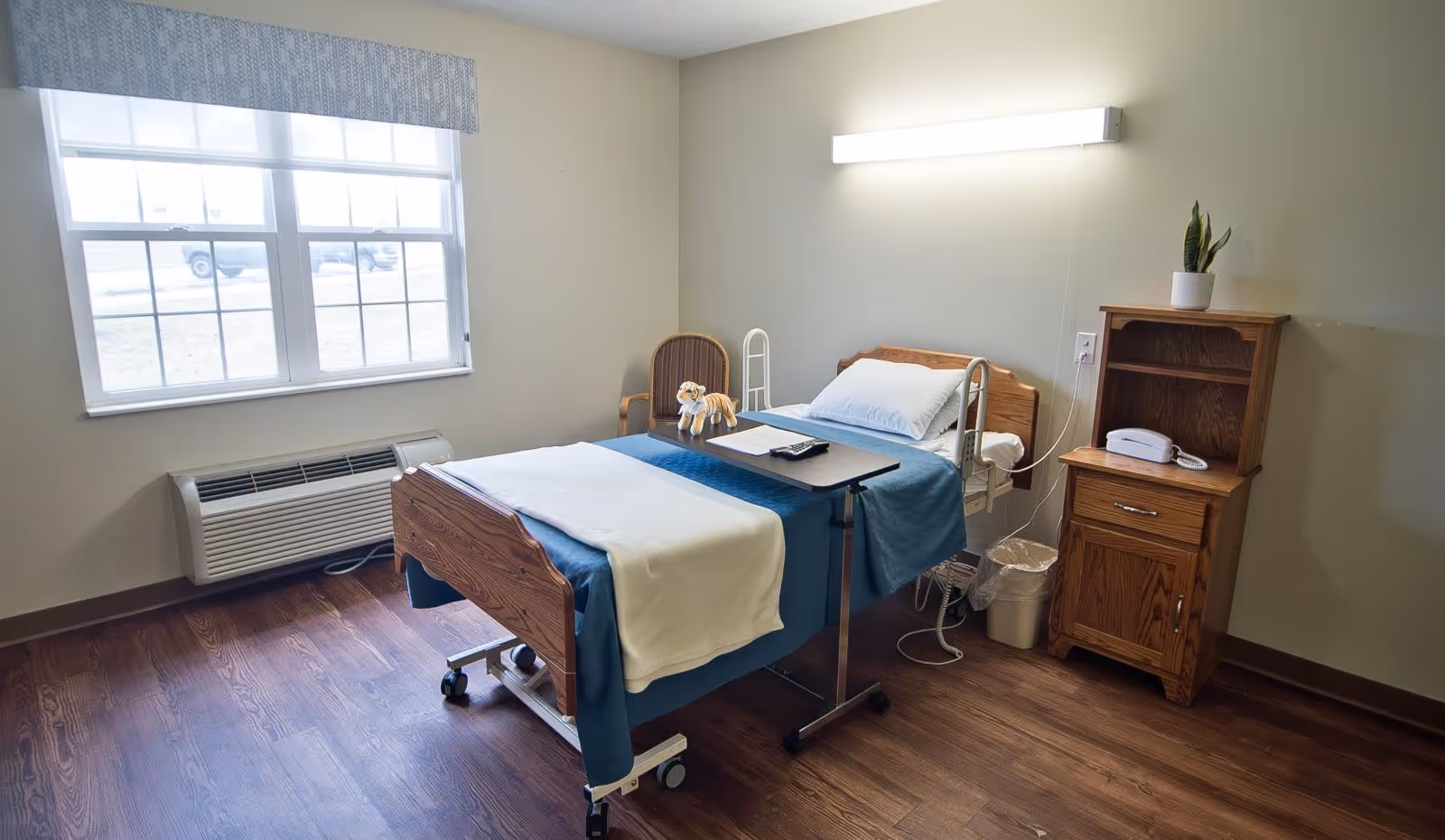 A simple, clean senior living facility bedroom with a hospital-style bed covered with blue and white bedding, a small overbed table with a remote control, a wooden nightstand with a white telephone and a small potted plant, a wooden chair with a stuffed animal on it, a window with blinds, and a wall-mounted light above the bed.