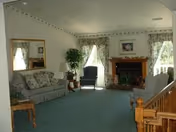 Bright living room with a floral-upholstered sofa and armchair, fireplace with a mantel, large mirror, and windows with floral curtains.
