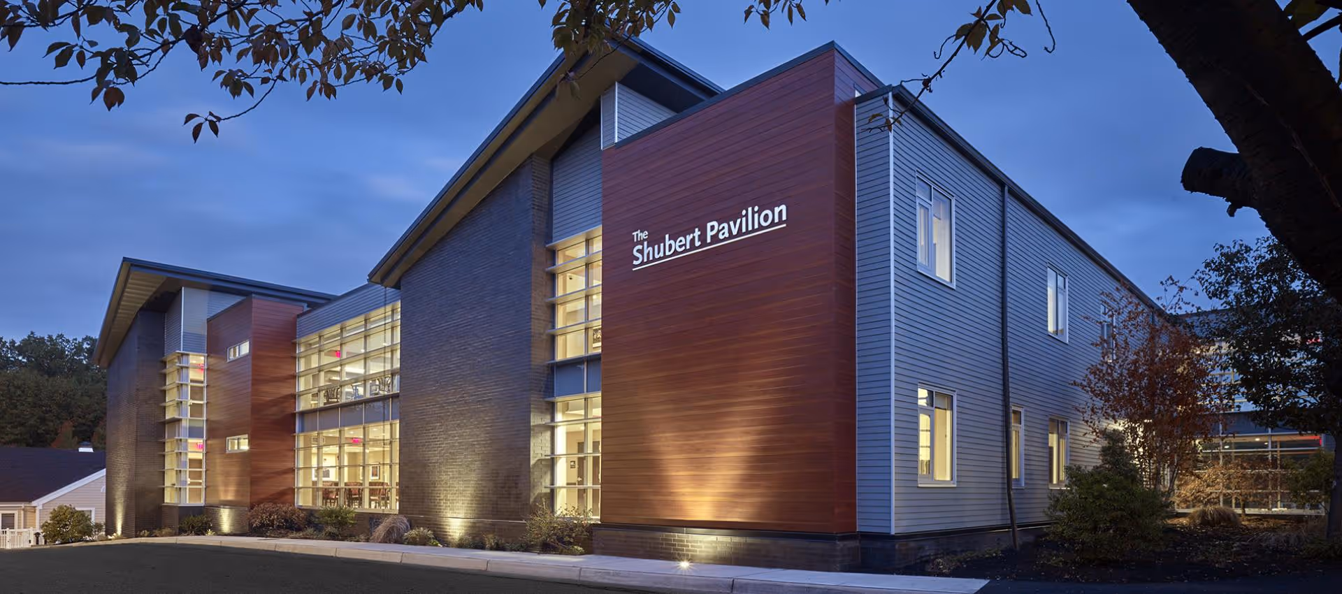 Exterior view of The Shubert Pavilion building at dusk, showing modern architecture with large windows, illuminated from inside, surrounded by trees and landscaping.