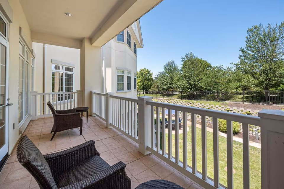Covered balcony with wicker chairs and a small table overlooking landscaped grounds and trees.