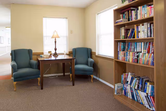 A cozy reading nook with two blue upholstered armchairs on either side of a wooden table with a lamp and books. To the right, there is a wooden bookshelf filled with various books. The room has beige walls and carpeted floor with two windows letting in natural light.