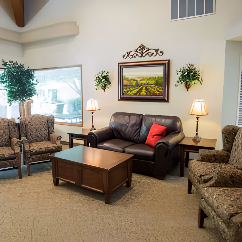 A cozy living room area with a dark leather loveseat featuring a red pillow, two patterned armchairs, a wooden coffee table, two side tables with lamps, and a framed landscape painting on the wall. There are also two wall-mounted plants and a large window letting in natural light.