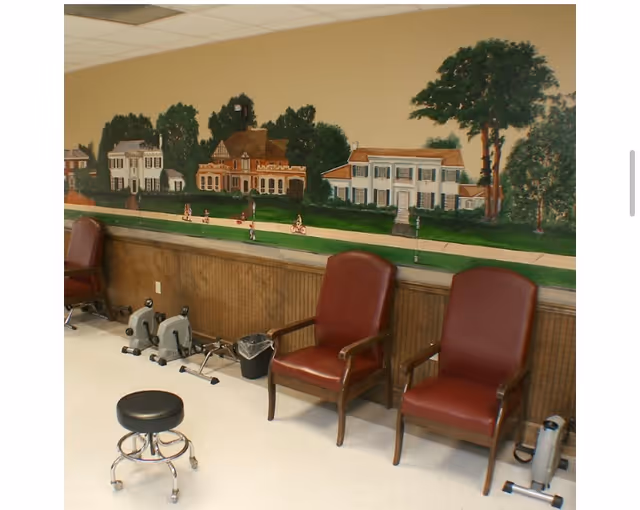 Room with two maroon armchairs and a black rolling stool on a white floor. There are two small exercise machines on the floor against the wall. The wall features a mural depicting houses, trees, and people walking and biking along a sidewalk.
