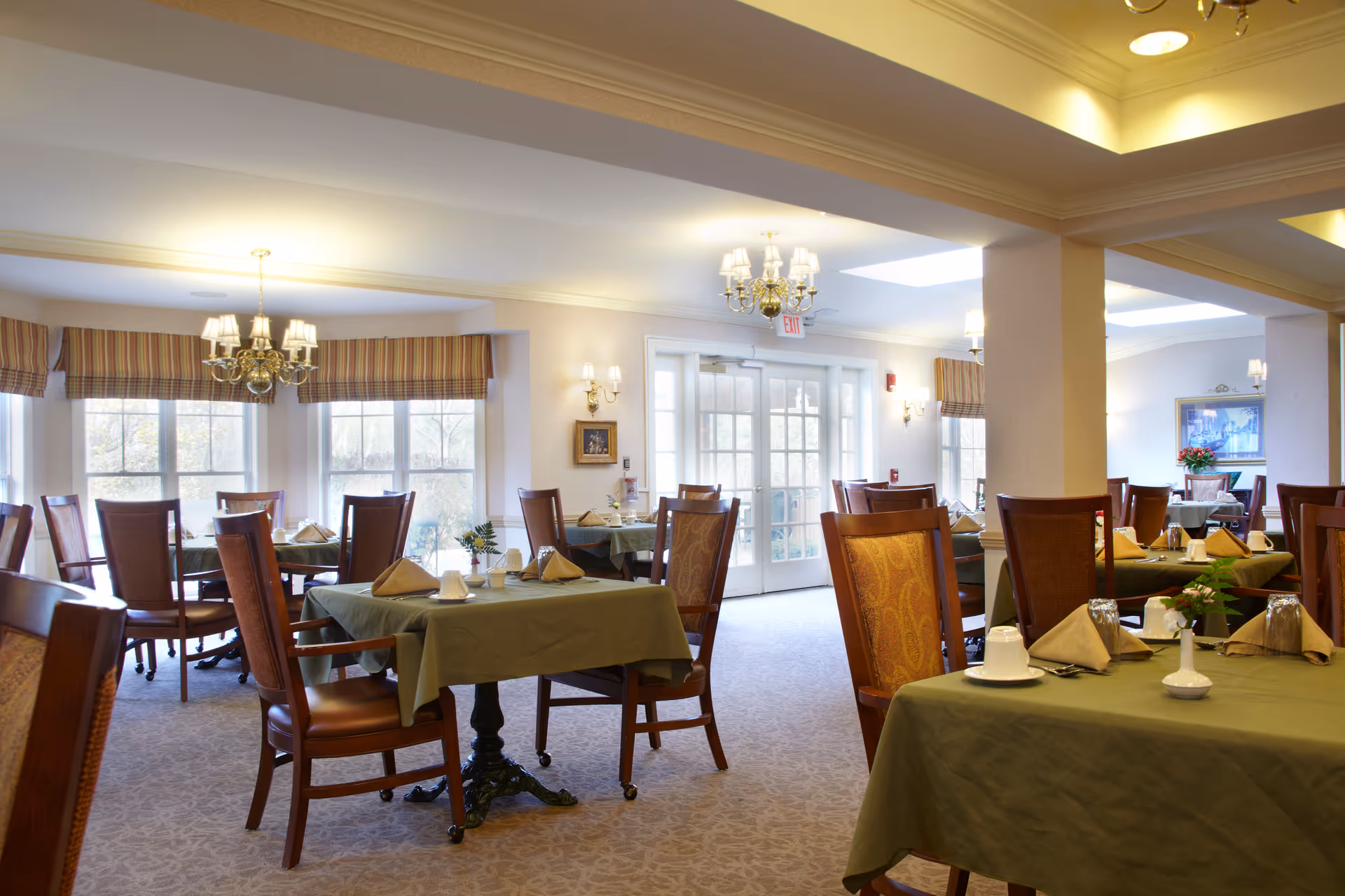 Bright dining room with multiple tables set with green tablecloths and wooden chairs under chandeliers.