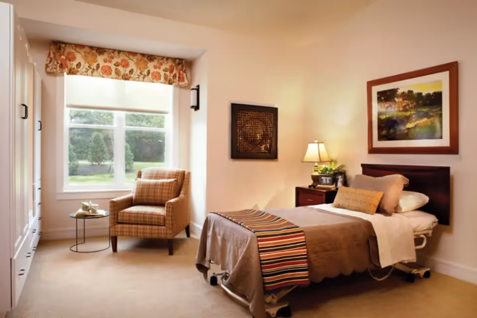 A cozy bedroom in a memory care facility featuring a single bed with brown bedding and a colorful striped throw, a wooden nightstand with a lamp and framed photos, a comfortable plaid armchair next to a small round side table, and a large window with floral valance letting in natural light. The walls are decorated with framed artwork.