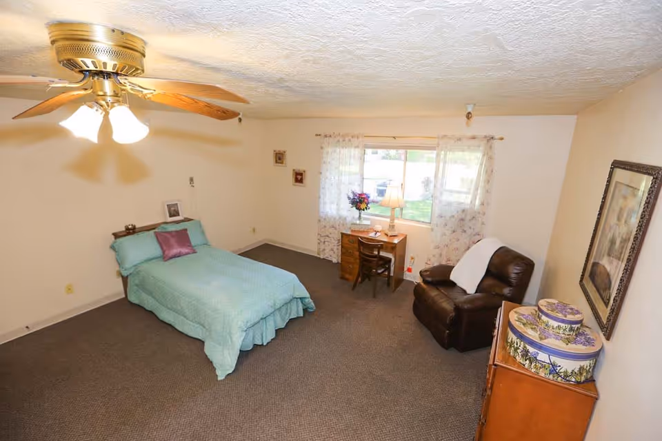 A roomy assisted-living bedroom with a single bed, desk by a window, leather recliner, ceiling fan and dresser.