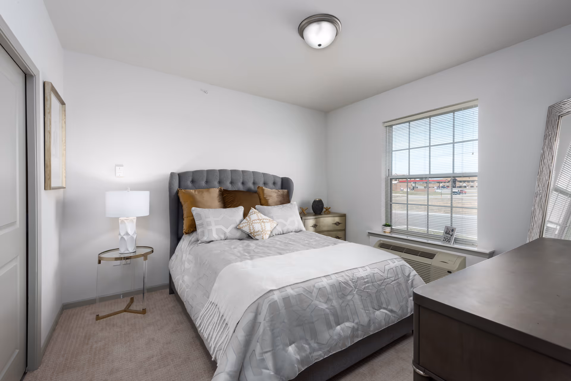 A neatly made bed with a tufted gray headboard and multiple pillows in a bright bedroom. The room has white walls, a window with blinds, a small side table with a lamp, a dresser, and a large mirror leaning against the wall.